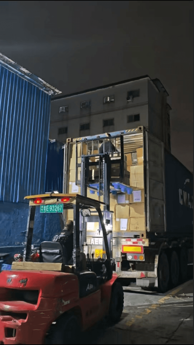 A forklift at the rear of an open shipping container containing stacked cartons at night.